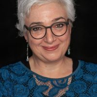 Portrait von Susan Renner-Haas. Sie hat graue Haare, trägte eine Brille mit schwarzem Rahmen und schaut lächelnd in die Kamera. Die trägte ein blaues Netzoberteil mit einer schwarzen Bluse darunter. Der Hintergrund ist schwarz-